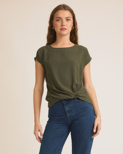 A woman modeling an olive green short-sleeve top with a twisted hem detail, paired with blue jeans, against a plain beige studio background.