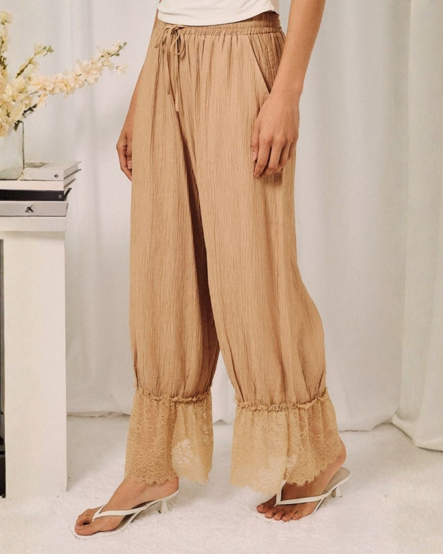 A person wearing loose beige pleated drawstring pants with ruffled hems, paired with a white top and white heeled sandals, standing in a softly lit indoor setting with a white curtain backdrop and a small side table with flowers.