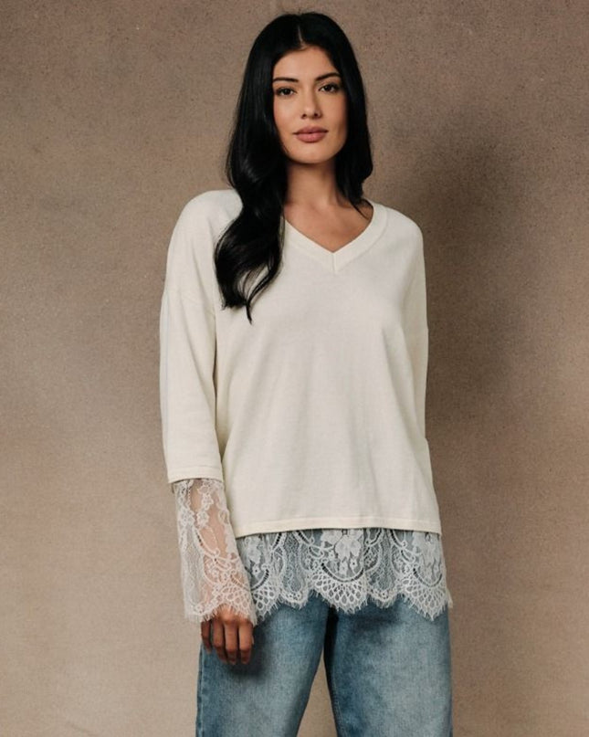 A woman modeling a cream-colored long-sleeve V-neck top with sheer lace bell sleeves and a matching lace hem, paired with light blue jeans, set against a textured beige studio backdrop.