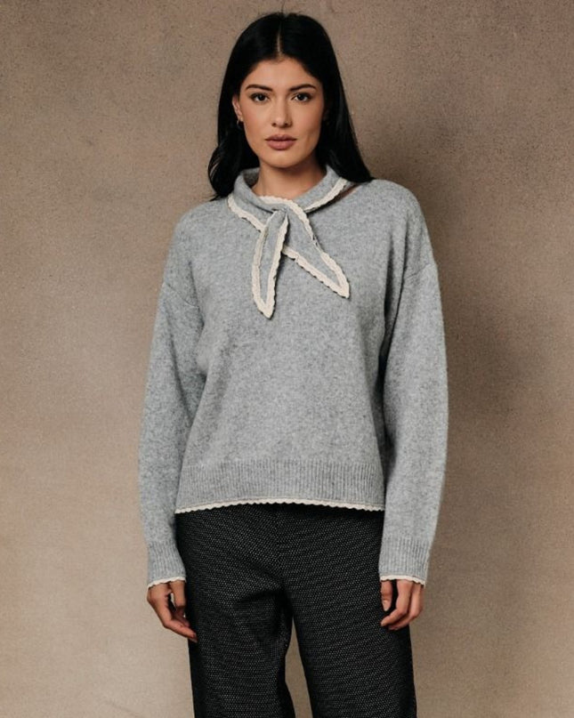 A woman modeling a light gray knit sweater with a cream-colored ruffled collar tie, paired with dark textured trousers, set against a neutral textured beige background.