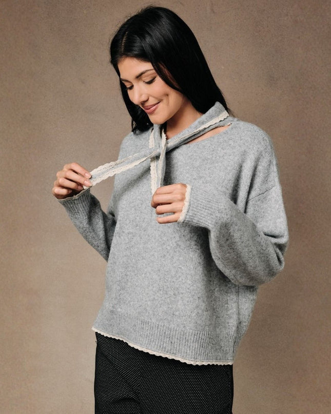 A woman wearing a light gray knit sweater with a unique cutout neckline and tie detail, paired with black pants, set against a plain brown studio background.