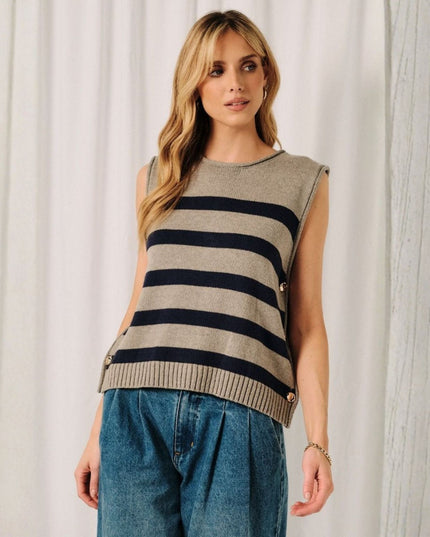 A woman wearing a sleeveless knit sweater vest with horizontal navy and beige stripes, styled with blue jeans against a white curtain backdrop.