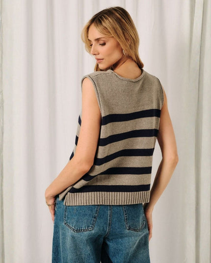 A woman wearing a sleeveless knit sweater vest with horizontal navy and beige stripes, paired with blue jeans, standing in front of a white curtain backdrop.