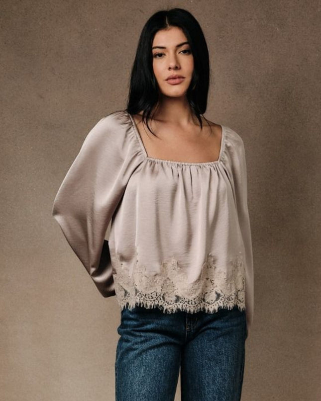 A woman modeling a light beige satin blouse with a square neckline, voluminous sleeves, and a lace hem, paired with blue jeans against a textured brown studio backdrop.
