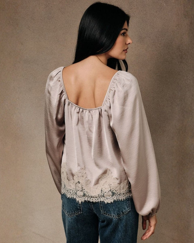 A woman shown from the back wearing a light taupe-beige long-sleeve blouse with a square neckline, gathered elastic trim, and lace detailing along the back hem, paired with blue jeans against a textured brown backdrop.