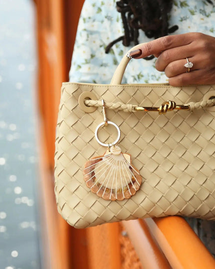 A close-up of a woven beige handbag with a top handle, gold-tone hardware, and a shell-shaped charm, held by a person wearing a light floral dress.