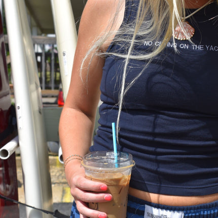 A close-up lifestyle shot of a woman wearing a navy blue ribbed tank top with white text that reads 'NO CLOSING ON THE YAC' and holding an iced coffee with a blue straw.