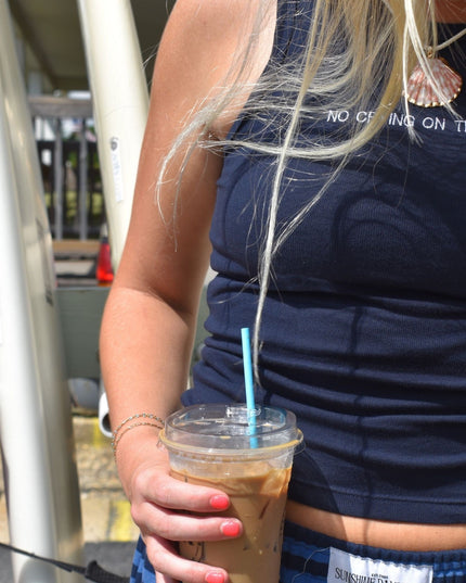 A close-up lifestyle shot of a woman wearing a navy blue ribbed tank top with white text that reads 'NO CLOSING ON THE YAC' and holding an iced coffee with a blue straw.