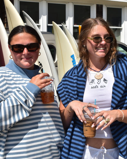Two women standing outdoors in front of surfboards and a building, each holding an iced coffee. The woman on the left wears a blue and white striped long-sleeve shirt, and the woman on the right wears a matching blue and white striped open-front cover-up over a white graphic T-shirt that reads 'salt air'.