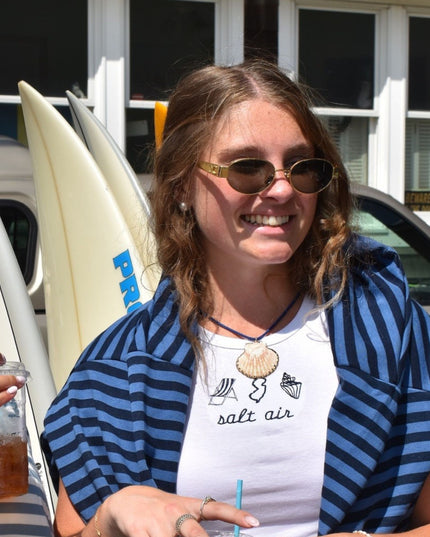 A smiling woman outdoors wearing gold-framed sunglasses, a blue and navy striped cardigan over a white graphic T-shirt that reads 'salt air,' with surfboards and a car visible in the background.