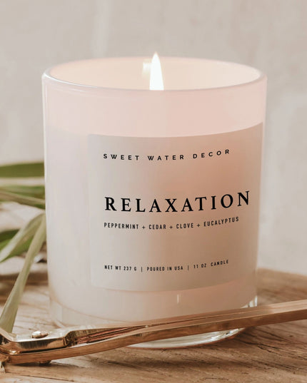 Candle labeled 'Relaxation' by Sweet Water Decor on a wooden surface with greenery.