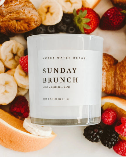 Candle labeled 'Sunday Brunch' surrounded by fruits and pastries on a white surface