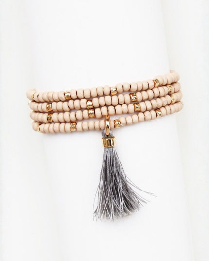 A multi-strand beaded bracelet with light beige wooden beads, small gold spacer beads, and a long gray tassel hanging from the center, displayed on a white mannequin arm.