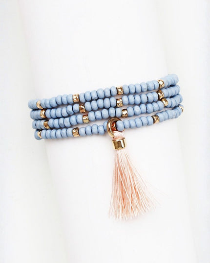 A multi-strand beaded wrap bracelet in light blue with small gold spacer beads and a dangling blush pink tassel, displayed on a white mannequin arm.