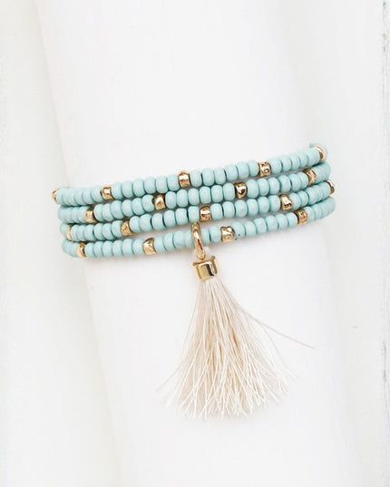 A multi-strand beaded bracelet with light blue beads, small gold spacer beads, and a cream tassel, displayed on a white cylindrical stand.