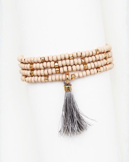 A multi-strand beaded wrap bracelet with light beige wooden beads, small gold spacer rings, and a dangling gray tassel with gold-toned hardware, displayed on a white mannequin arm.