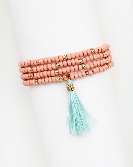 A multi-strand beaded wrap bracelet in a dusty pink color, accented with small gold spacer beads and a dangling mint green tassel charm, displayed on a white cylindrical mannequin arm.