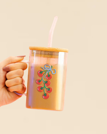 Hand holding a clear tumbler with a bamboo lid and straw, featuring a design of red tomatoes and green leaves on a beige background.