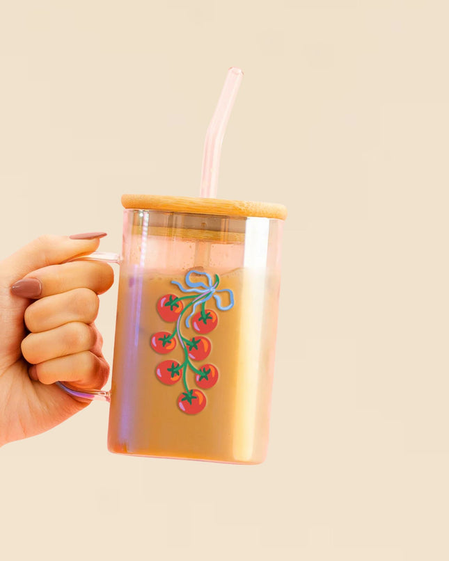 Hand holding a clear tumbler with a bamboo lid and straw, featuring a design of red tomatoes and green leaves on a beige background.