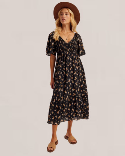 A woman modeling a black midi dress with a small beige floral pattern, styled with a brown wide-brim hat and brown slide sandals against a light beige studio background.
