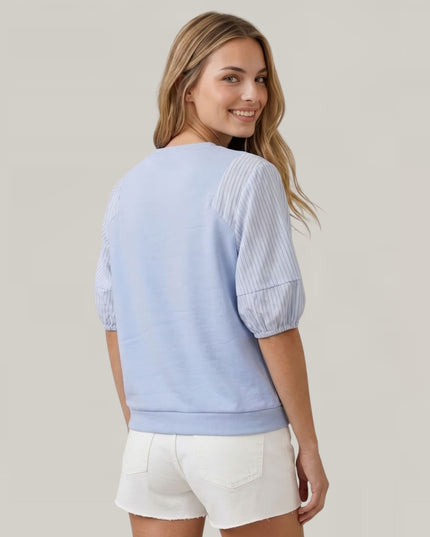 A woman shown from the back wearing a light blue short-sleeve top with textured vertical stripes on the sleeves and white shorts, against a plain light gray studio background.